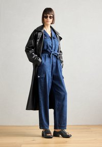 Salopette in denim blu scuro con maniche corte, vita alta con cintura e tasche, indossata sotto un cappotto in pelle nera. Zoccoli neri aperti.