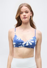 Νεαρή γυναίκα με μακριά ξανθά μαλλιά, φορώντας μπλε και λευκό φλοράλ κοντό τοπ και ψηλόμεσα λευκά σορτς, στέκεται μπροστά σε απλό φόντο.