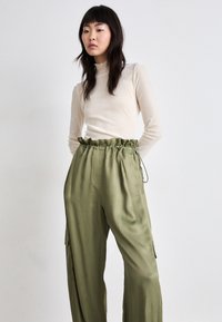 Cream long-sleeve top with a ruffled neckline paired with loose, olive green satin cargo pants featuring side pockets and an adjustable waistband.