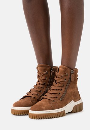 Bottines en suède marron avec une semelle blanche épaisse, comportant des accents de fermeture éclair en métal et un laçage à l'avant. Finition texturée et design montante.