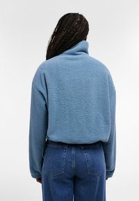 Personne aux cheveux tressés portant une veste polaire bleu clair et un jean bleu, vue de dos sur un fond uni.