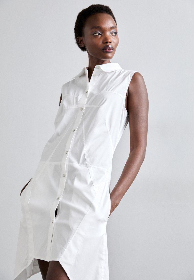 Hvid ærmeløs kjole med knapfront, spidse krave og sidelommer. Har en glat tekstur og strukturerede sømme for en skræddersyet pasform.