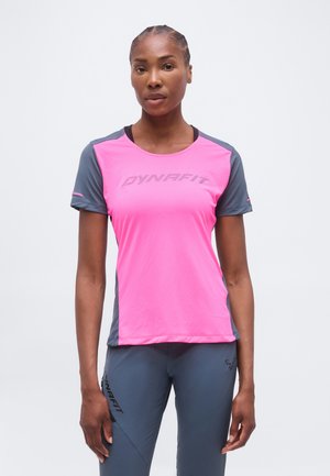 Γυναίκα που στέκεται αντικρίζοντας μπροστά, φοράει ροζ και γκρι αθλητική μπλούζα με κοντά μανίκια και γκρι αθλητικό παντελόνι με λογότυπα.