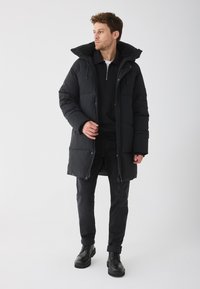 Sort pufferjakke med hætte, med lynlås foran, lange ærmer og lommer. Båret over en sort sweater og bukser med sorte støvler.