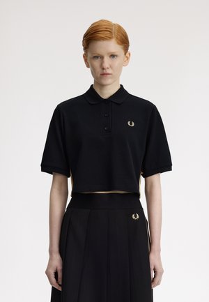 Personne aux cheveux roux portant un polo court noir et une jupe plissée taille haute noire, tous deux ornés de petits logos en forme de couronne de laurier dorée.