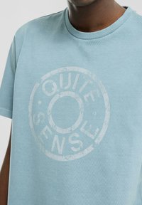 T-shirt en coton bleu clair avec un graphisme circulaire usé affichant les mots « QUIT » et « SENSE » imprimés en blanc. Manches courtes, col rond.