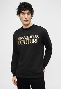 Νεαρός άνδρας με σγουρά σκουρόχρωμα μαλλιά, φοράει μαύρο φούτερ με χρυσά γράμματα «Versace Jeans Couture» και στέκεται μπροστά σε έναν απλό φόντο.