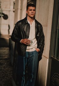 Black leather jacket, white knit polo, and blue denim jeans. Holding a white coffee cup, standing against a stone wall in soft sunlight.