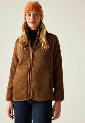 Femme portant une veste polaire marron à fermeture éclair, un bonnet en tricot orange, un col roulé noir et un jean bleu foncé sur un fond uni.
