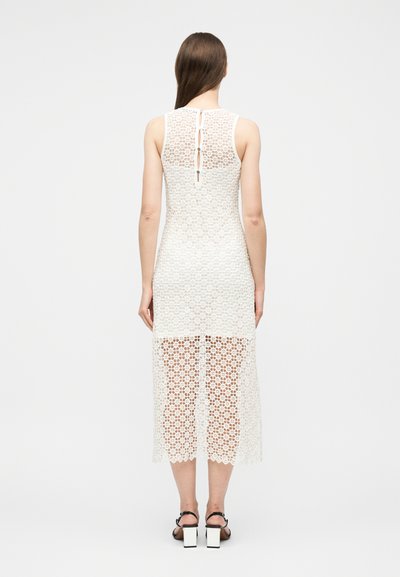 Femme aux longs cheveux bruns portant une robe midi blanche en dentelle sans manches avec des boutons en forme de trou de serrure dans le dos, associée à des talons blocs noirs à bride.