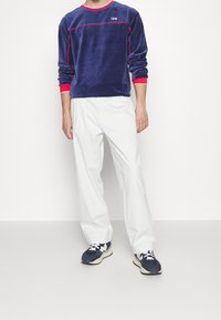 Námorně modrá sametová košile s dlouhým rukávem, červenými akcenty a logem, kombinovaná s bílými volnými kalhotami a námorně modrými sportovními teniskami.