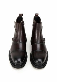 Botas de tobillo de cuero marrón con un acabado suave, que presentan una punta redondeada, cierre de cremallera y detalles decorativos de hebilla. Suela de goma.