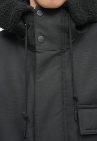 Must have mustla, millel on tekstuuriga, fliisvoodriga kapuuts, nööbid, reguleeritavad tõmblukud ja küljetasku. Sujuv välimine kangas.