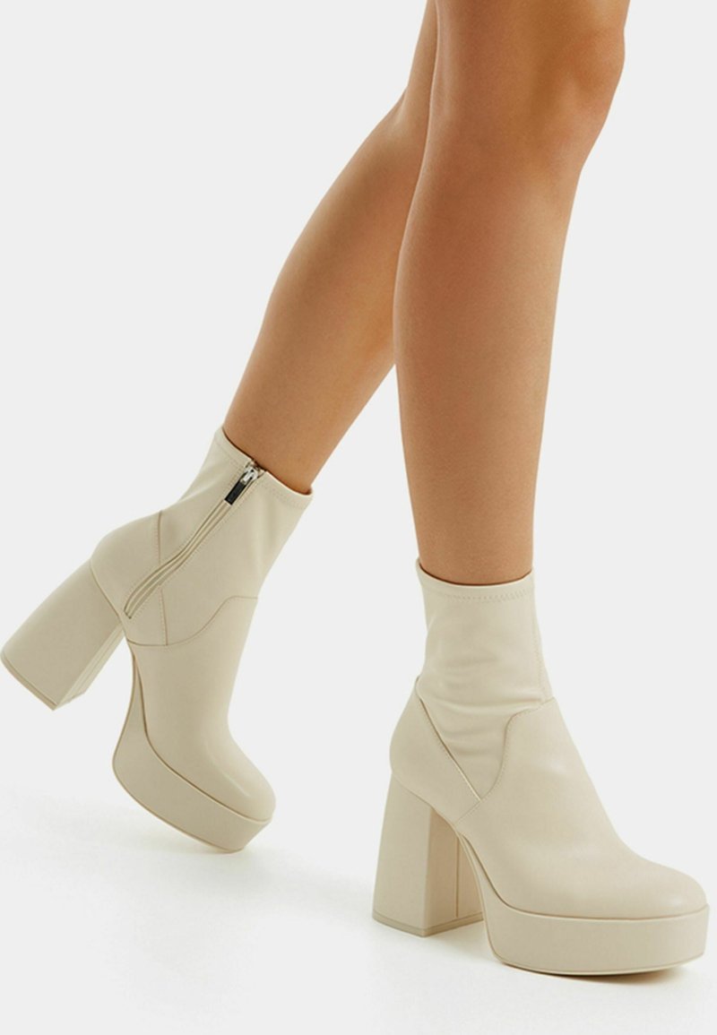 Jambes portant des bottines à plateforme couleur crème avec talons hauts et épais et fermetures éclair latérales sur fond blanc.