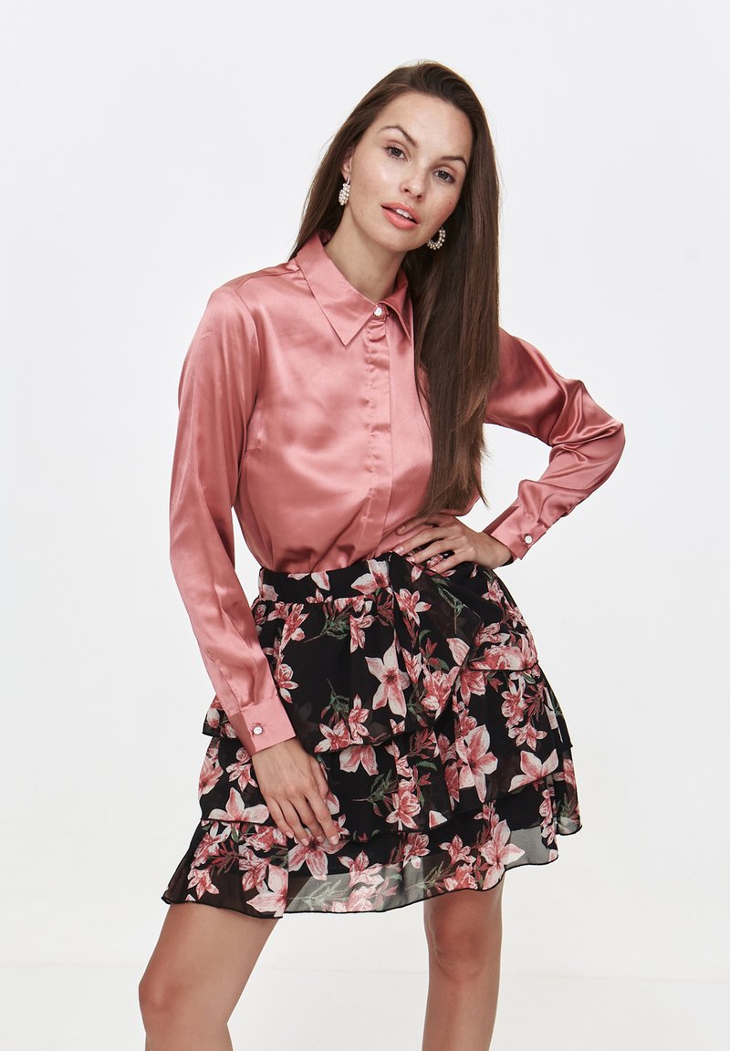 Różowa satynowa bluzka z długim rękawem i kołnierzykiem, połączona z czarną warstwową spódnicą w kwiaty z różowymi kwiatami. Gładka tekstura i dopasowany fason.