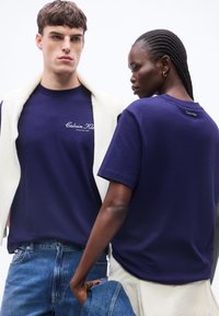 Δύο μοντέλα φοράνε μπλε ναυτικό T-shirts της Calvin Klein, το ένα κοιτάει μπροστά με ένα λευκό πουλόβερ ριγμένο στους ώμους, το άλλο δείχνει την πλάτη.