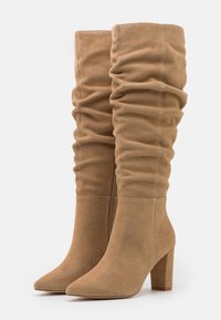 Bottes hauteur genou, décontractées, en daim beige, avec un bout pointu et un talon bloc ; présentent des coutures apparentes et une texture lisse.