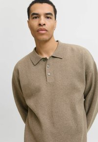 Mladić s kratkom kovrčavom kosom nosi bež dugu majicu s dugim rukavima i stoji protiv jednostavne svijetle pozadine.