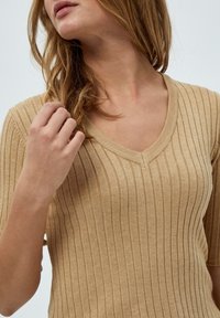 Top en tricot beige à côtes avec un décolleté en V et des manches courtes. Le tissu a une texture douce et de légères rayures verticales.