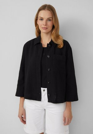 Zwarte cropped shirtjas van textuurstof, met knopen aan de voorkant en een borstzak, gecombineerd met een zwarte top en witte shorts.