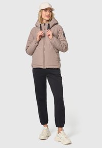 Pufferjakke i lys brun med hætte, frontlynlås, to sidelommer og sorte snore. Samparet med mørke joggers og sneakers.