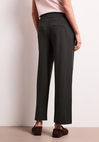 Femme portant un pantalon noir droit avec des poches passepoilées à l'arrière, une chemise rose pâle rentrée et des mocassins marron, debout contre un mur clair.