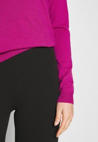 Fuchsia gestrickter Pullover mit gerippten Bündchen über schwarzer, taillierter Hose, die glatte Texturen und schlichtes Design zeigt. Hand auf der Taille ruhend.