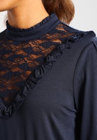 Marinblus med spetspanel och volangkrage. Mjuk tyg med en texturerad spets överdrag, som framhäver intrikata mönster.