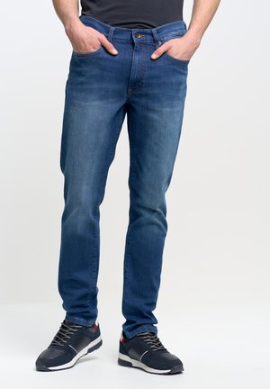 Temno modri jeans s slim fit krojem, ki imajo pet žepov in gumbasto zapiranje. V kombinaciji z črnimi športnimi čevlji z rdečimi detajli.