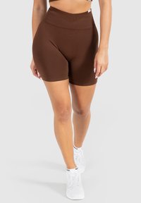 Bruine high-waisted shorts van rekbaar materiaal, met een gladde textuur en een getailleerd ontwerp, aangevuld met witte sneakers.