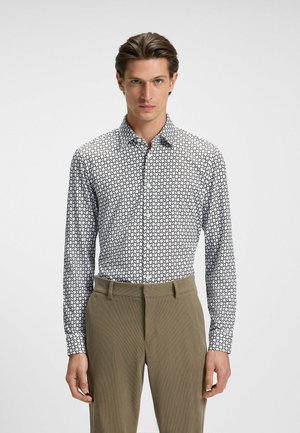 Chemise blanche à manches longues avec un motif géométrique noir, col boutonné et coupe ajustée, associée à un pantalon léger beige.