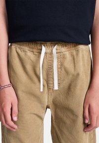 Enfant portant un pantalon en velours côtelé beige avec un cordon blanc et un haut noir, les mains relâchées le long du corps, un poignet orné d'un bracelet rouge tissé.