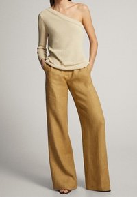 Top a coste beige a spalla unica, abbinato a pantaloni ampi color marrone chiaro, caratterizzati da una texture morbida e una vestibilità comoda.