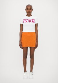 Fehér, testhezálló póló rózsaszín "VERSACE JEANS COUTURE" felirattal, élénk narancssárga, gyöngyökkel díszített mini szoknyával és fehér sportcipőkkel párosítva.