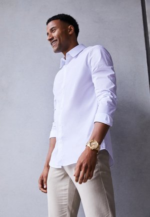 Chemise blanche à manches longues avec boutons, associée à un pantalon beige. Montre en or au poignet, tissu lisse, coupe ajustée et manches retroussées.