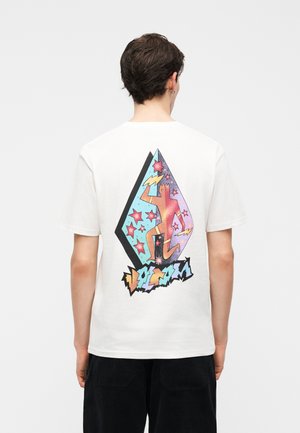 Νέος άνδρας με κοντά μαλλιά που φοράει λευκό μπλουζάκι με πολύχρωμη αφηρημένη φιγούρα, αστέρια και κεραυνούς στη πλάτη.