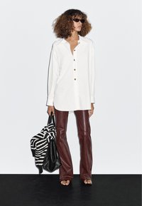 Bílá košile na knoflíky s volnějším střihem, hnědé zvrásněné kalhoty z umělé kůže, černá kabelka a pruhovaný svetr, nošeno s černými slunečními brýlemi.