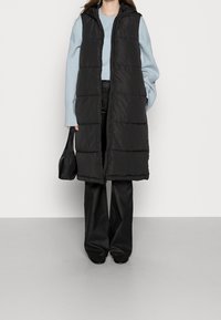 Czarna pikowana kamizelka z kapturem, z poziomym wzorem pikowania, zestawiona z jasnoniebieskim swetrem i czarnymi szerokimi spodniami. Czarna torebka.