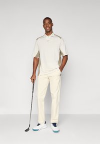 Men's golf outfit featuring a beige polo shirt with gray accents and cream trousers. Includes a golf club and white shoes with blue details.