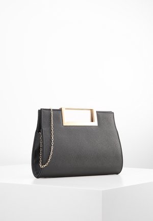Schwarze strukturierte Handtasche mit rechteckigem goldfarbenem Metallgriff und dünnem goldfarbenem Kettenriemen, der auf einer weißen Oberfläche liegt.