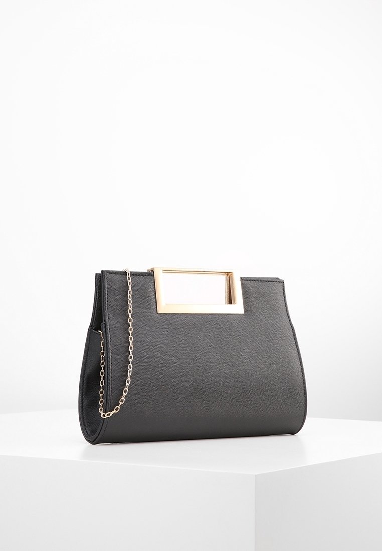 Sac à main noir texturé avec une poignée rectangulaire en métal doré et une fine bandoulière chaîne dorée reposant sur une surface blanche.