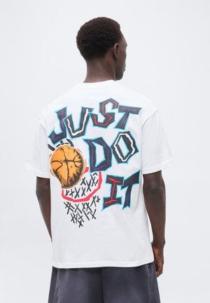 Homme portant un T-shirt blanc avec un texte coloré style graffiti « JUST DO IT » et un graphisme de ballon de basket orange dans le dos.