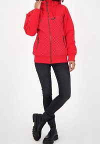 Röd zip-up jacka med prickigt mönster, med huva och två svarta dragkedjefickor. Bärs med mörka jeans och svarta ankelstövlar.