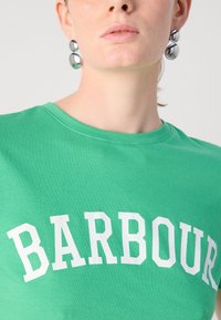Zelené bavlněné tričko s výrazným bílým nápisem "BARBOUR." Kulatý výstřih, měkká textura a krátké rukávy.