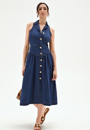 Femme portant une robe en jean bleu foncé sans manches, boutonnée avec col, tenant un sac tressé marron, debout devant un fond uni.