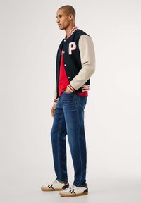 Moška jakna v stilu varsity v mornarsko modri barvi z kremnimi rokavi, rdečimi poudarki in črko P. Združena z rdečo majico, modrimi kavbojkami in belimi supergami.