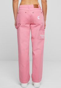 Pantalones cargo de denim rosa con un corte relajado, que cuentan con bolsillos traseros y detalles de marca bordados. Unas zapatillas blancas lisas contrastan con el color.