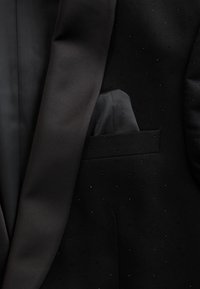 Chaqueta de esmoquin negra con solapa de satén y un pañuelo negro guardado en el bolsillo del pecho, con sutiles puntos texturizados en la tela.