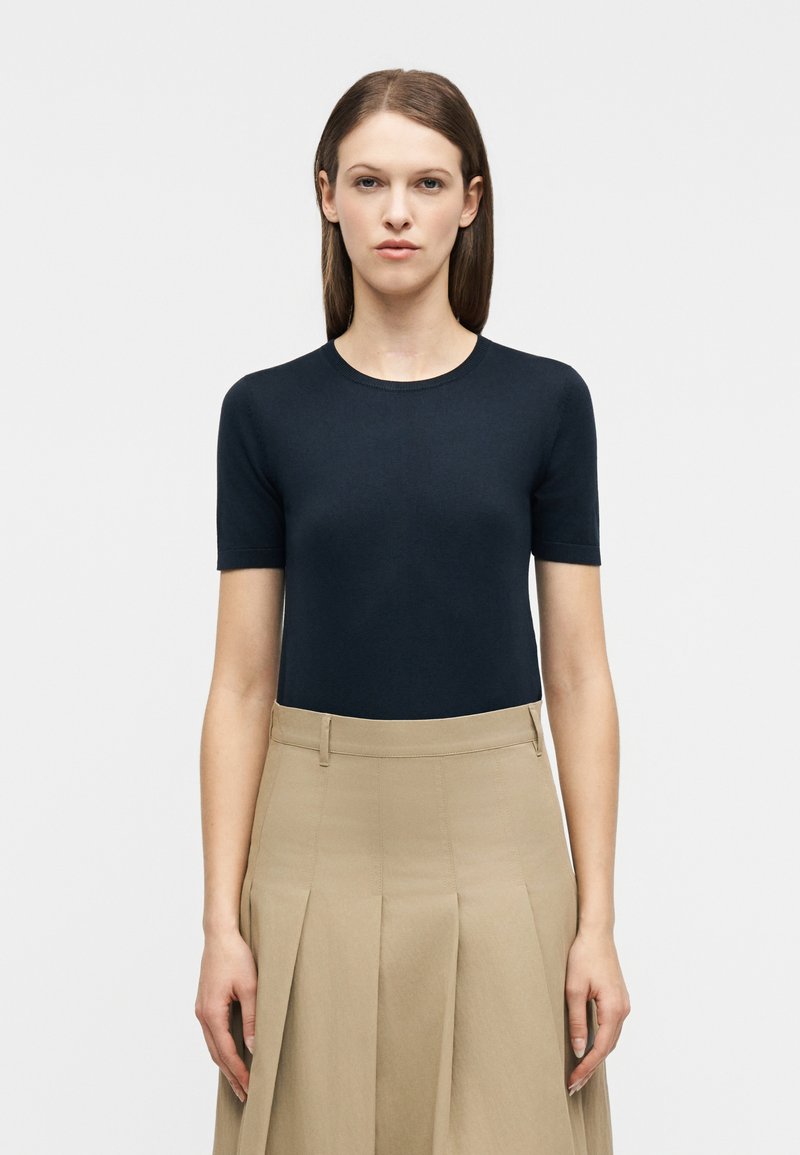 Γυναίκα με ίσια καστανά μαλλιά, φοράει μια εφαρμοστή μπλε ναυτική κοντομάνικη μπλούζα που είναι μπαλωμένη σε μια ψηλόμεση μπεζ πλισέ φούστα.