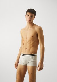 Menns beige bomullsboksere med svart elastisk midjebånd som har en logo. Designet inkluderer en støttende frontlomme og en tettsittende fasong.